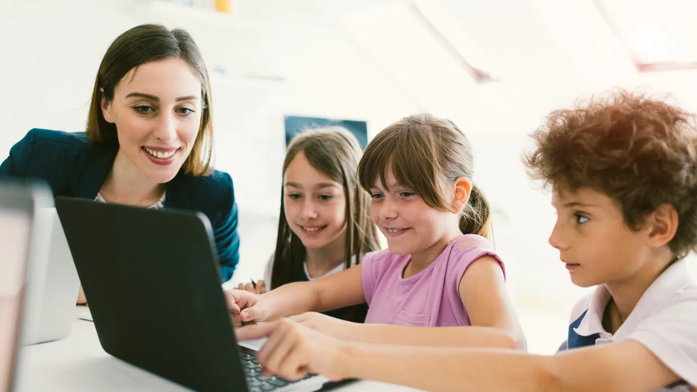 An image of a teacher observing 3 children, who are looking at a laptop and viewing the 25 best scratch games to play.